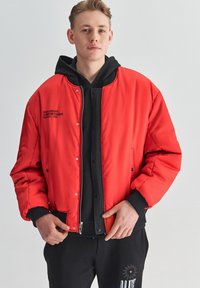 Czerwona ocieplana kurtka bomberka z ribbowymi mankietami i kołnierzem, z czarnym zamkiem błyskawicznym i zapięciem na zatrzask oraz wytłoczonym logo na klatce piersiowej.