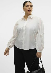 Biała bluzka zapinana na guziki z długimi, objętościowymi rękawami i wyszywanymi detalami na mankietach. W połączeniu z czarnymi spodniami i czarną torebką.