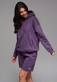 Sweatshirt à capuche violet avec des manches longues et une poche avant, associé à un short violet assorti. Tissu doux, design décontracté, avec un détail de logo.