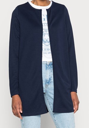 Persoon draagt een marineblauwe openvallende cardigan met lange mouwen over een witte T-shirt met print en lichtblauwe jeans.