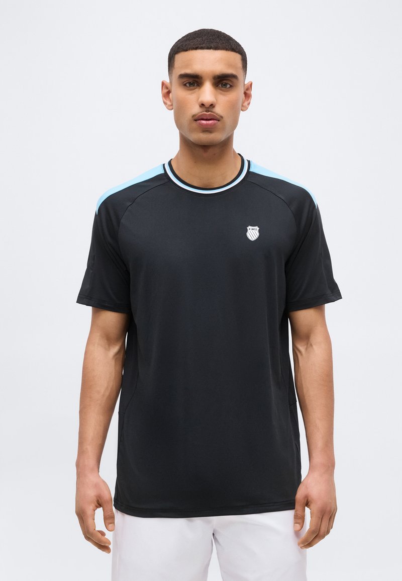 Άνδρας που φοράει μαύρο αθλητικό T-shirt με ανοιχτό μπλε ρίγες στους ώμους και ένα μικρό έμβλημα με σχήμα ασπίδας στο αριστερό στήθος, στέκεται μπροστά σε απλό φόντο.