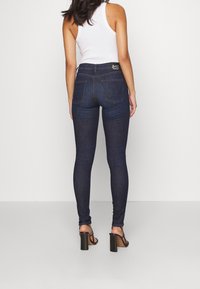 Vaqueros skinny de color azul oscuro con tiro alto, dos bolsillos traseros y detalles de costura sutiles, combinados con sandalias de tacón negro.