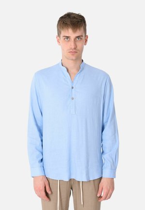 Giovane uomo che guarda avanti, indossa una camicia azzurro chiaro a maniche lunghe con una pattina parziale di bottoni e pantaloni beige con coulisse.