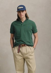 Jaunas vyras stovi su rankomis kišenėse, vilkintis žalią polo marškinėlį, smėlio spalvos kelnes su dryžuotu diržu, mėlyną "Ralph" kepurę ir laikrodį kairiajame rieše.
