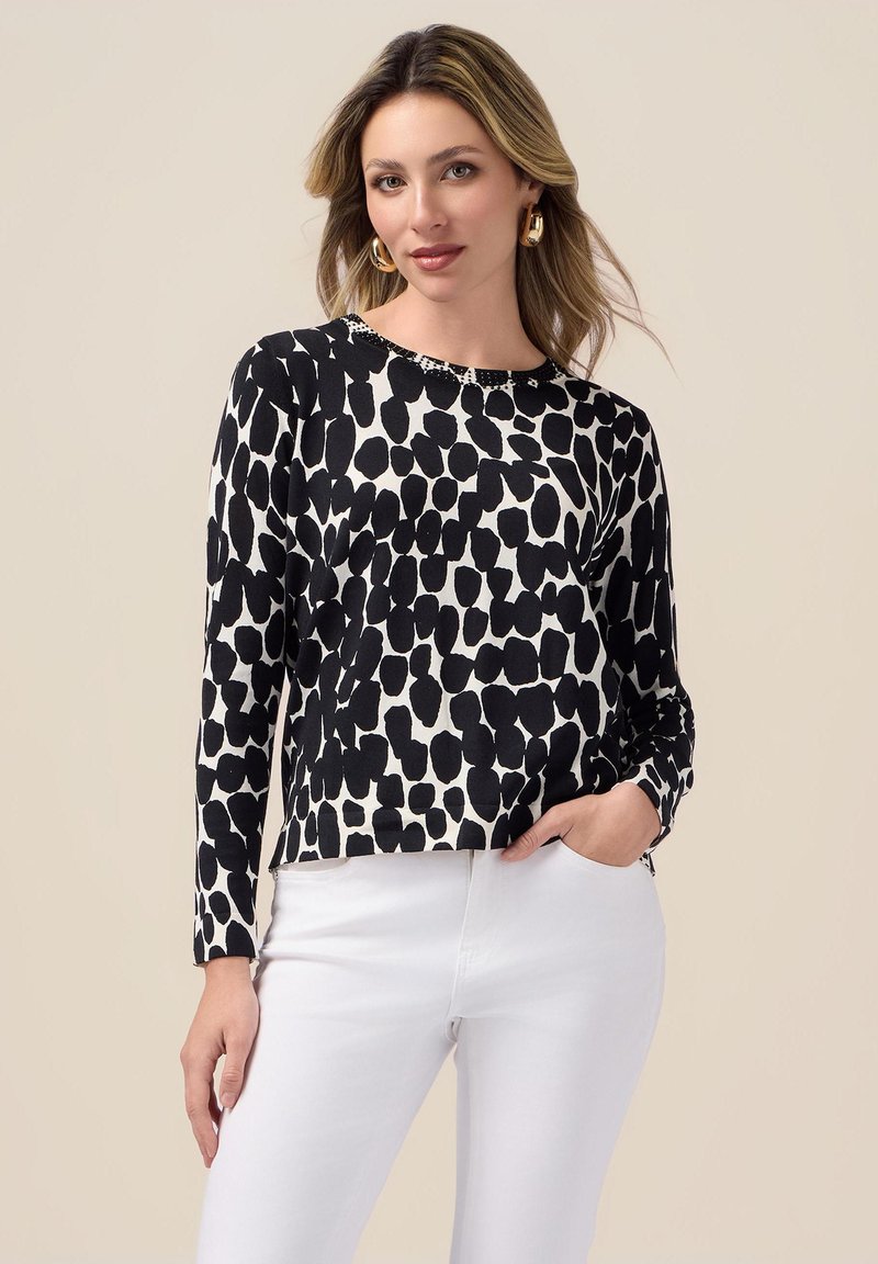 Cămașă pe mânecă lungă alb-negru, cu un model abstract, guler rotund și o croială lejeră, purtată cu pantaloni albi.