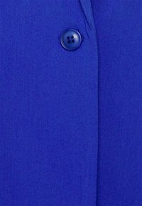 Tissu bleu royal à la texture lisse ; présente un bouton rond à quatre trous et un détail de couture subtil le long de la couture.