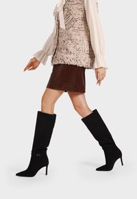 Stivali alti in suede nero con punta affilata e tacchi a stiletto, dotati di una sottile cinghia per la caviglia per un tocco in più. Texture liscia in generale.
