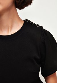 Top nero a maniche corte con scollatura rotonda, caratterizzato da tre accenti circolari in metallo sulla spalla e un tessuto liscio e strutturato.