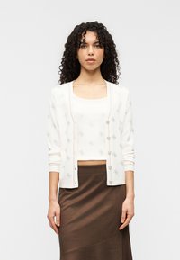 Λευκό ζακέτα με γκρι διακοσμητικές λεπτομέρειες, διαθέτει κουμπιά μπροστά και μακριά μανίκια, συνδυασμένο με κρεμ τοπ και καφέ φούστα.