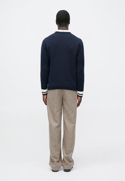 Άνδρας με πλάτη, φοράει μπλε πουλόβερ με λευκές ρίγες στα μανίκια και μπεζ παντελόνι φαρδιάς γραμμής, στέκεται μπροστά σε απλό λευκό φόντο.