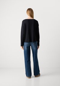 Woman with long straight hair stands facing away, wearing a black long-sleeve top, wide-leg blue jeans, and brown shoes against a plain background.