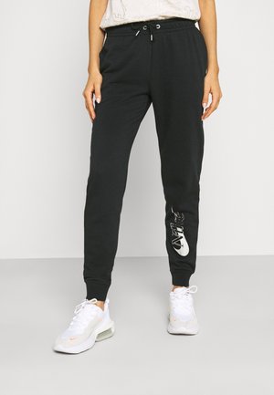 Persoon draagt zwarte Nike joggingbroek met elastische tailleband en witte sneakers, staand tegen een effen achtergrond.
