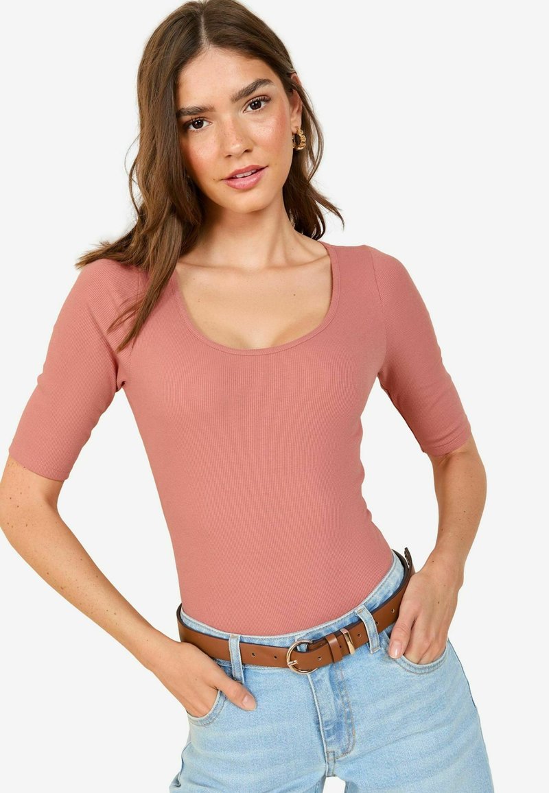 Kerge roosa ribidega bodi lühikeste varrukatega ja ümar kaelus, koos siniste kõrge vöökohaga teksade ja pruuni nahkvööga.