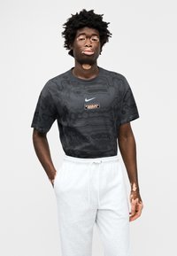 Μαύρο T-shirt με κοντά μανίκια και υφασμένο μοτίβο, με λευκό λογότυπο Nike και ορθογώνια πορτοκαλί ετικέτα, συνδυασμένο με γκρι φόρμες.