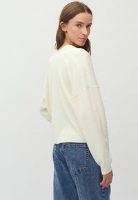 Pulover crem cu o croială relaxată, umeri lăsați și manșete cu nervuri. Asortat cu blugi lejeri albaștri. Textură netedă și design simplu.