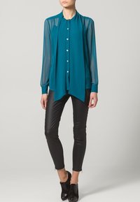 Blusa teal con maniche trasparenti, frontale con bottoni e design fluido. Abbinata a pantaloni neri aderenti e scarpe stringate.