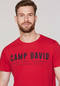 Red cotton t-shirt featuring a crew neck and short sleeves. Text printed in black: "CAMP DAVID REGISTERED TRADEMARK AUTHENTIC STYLE."