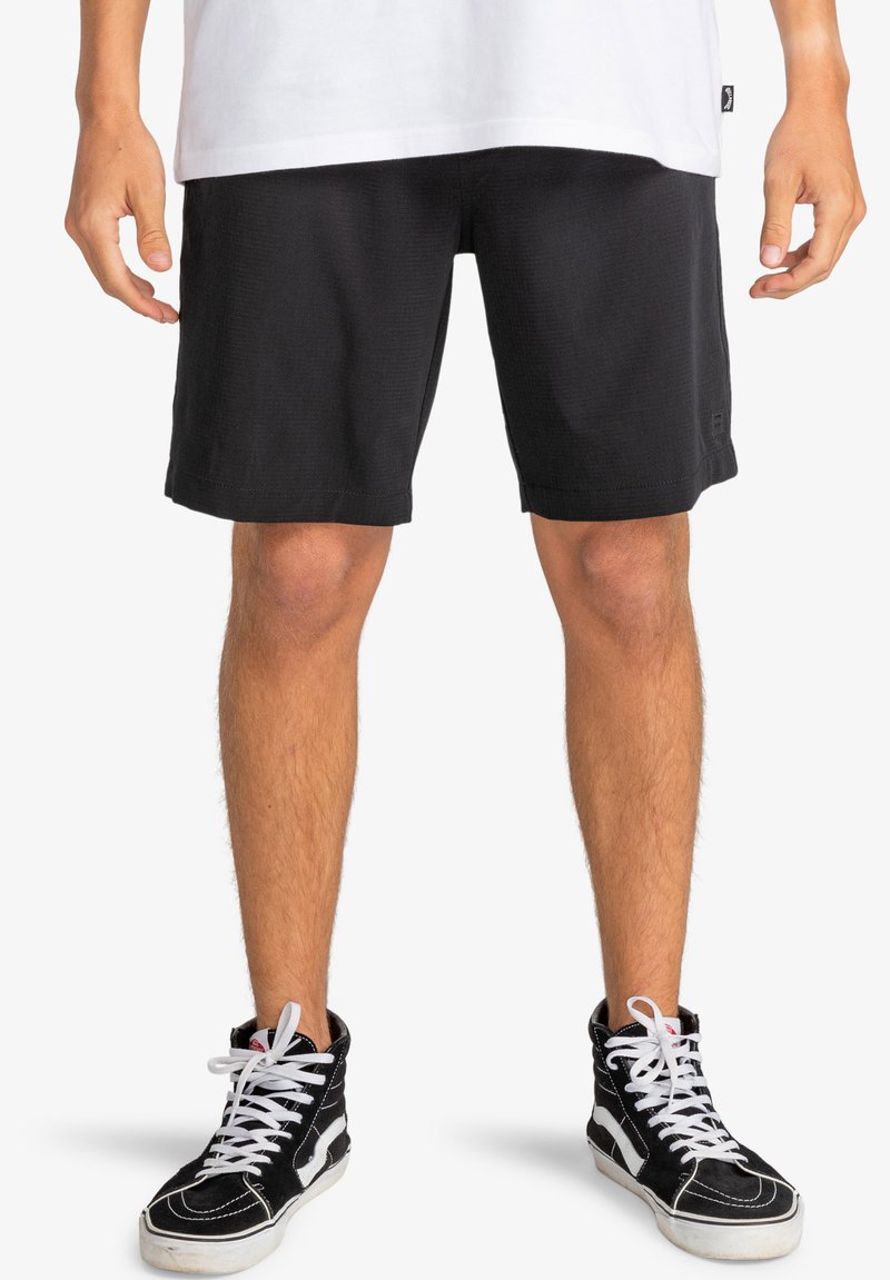 Pantalones cortos negros de tela texturada, con un ajuste recto y dobladillo. Combinados con zapatillas negras de caña alta con acentos blancos.