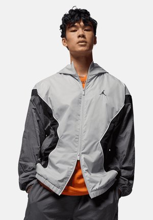 Joven viste chaqueta con cremallera gris y negra con pequeño logo en el pecho sobre camisa naranja, de pie con las manos en los bolsillos.