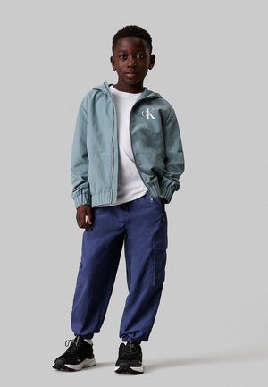 Chaqueta con capucha de color azul claro, con puños elásticos y un logo, combinada con pantalones cargo de color navy y zapatillas deportivas negras. Camiseta blanca por debajo.