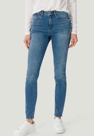 Femme portant un jean bleu taille moyenne slim, associé à des baskets blanches et un haut blanc à manches longues, debout devant un fond uni.