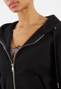 Čierna mikina na zips s fleecovou podšívkou, strieborný zips, kapucňa na šnúrky a leopardí vzor, ktorý vykúka spodnej časti.