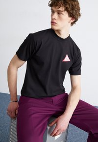 Camiseta negra de manga corta con un logo triangular rojo en el pecho, combinada con pantalones morados, sentada sobre una superficie metálica.