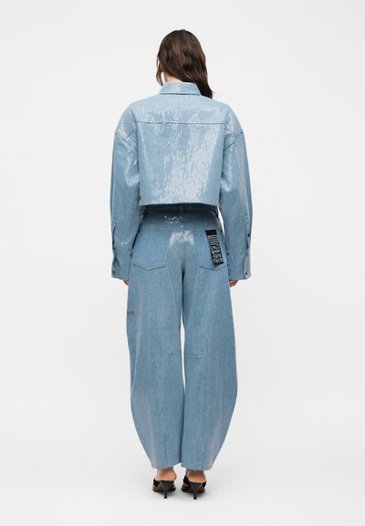 Γυναίκα που φοράει ένα ανοιχτό μπλε κροπ μπουφάν με παγιέτες και αντίστοιχο φαρδύ παντελόνι με μαύρο μπαλώμα, όρθια με την πλάτη γυρισμένη.