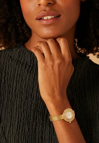 Gold mesh wristwatch with an oval face featuring a white dial, surrounded by decorative stones and textured fabric top.