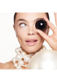 Mujer con collar de perlas sostiene una esfera negra sobre un ojo, mirando hacia arriba, con una gran perla blanca en primer plano.