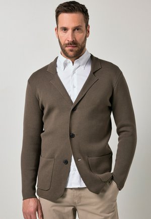 Mand iført brun knappet cardigan over hvid skjorte og beige bukser med den ene hånd i lommen mod en ensfarvet baggrund.