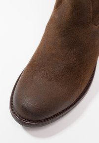 Bottine en daim marron avec un bout arrondi, bord sombre contrasté et une finition lisse. Présente des coutures minimales et un design épuré.