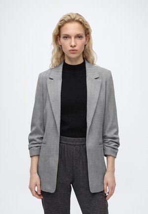 Femme aux cheveux blonds porte un blazer gris sur un top noir et un pantalon gris foncé, debout devant un fond blanc uni.