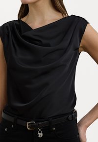 Zwarte satijnen top met gedrapeerde halslijn en mouwloos ontwerp. Draagt een zwarte riem met een zilveren gesp en decoratieve hangslotdetail.
