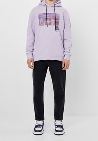 Sudadera ligero morado con un estampado gráfico, bolsillo frontal tipo canguro y cordones negros. Combinada con jeans negros y zapatillas multicolores.