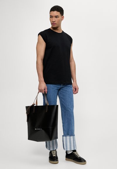 Borsa tote nera in pelle con un design strutturato, con manici in pelle marrone e un logo sulla parte frontale, abbinata a jeans blu e una maglietta nera.