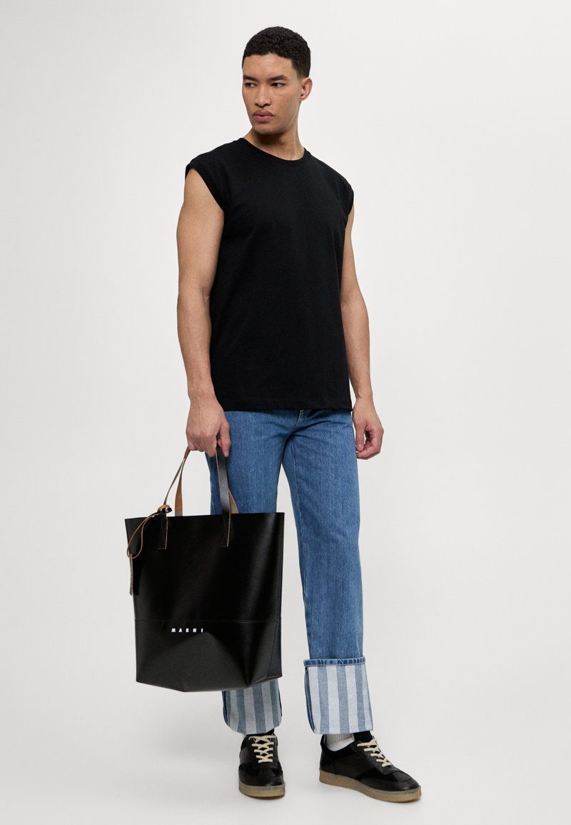 Sac fourre-tout en cuir noir au design structuré, avec des poignées en cuir marron et un logo à l'avant, associé à un jean bleu et une chemise noire.