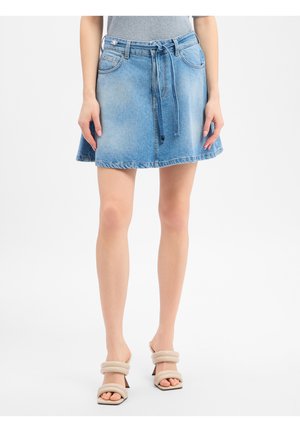 Frau trägt hellblauen Jeansrock mit Bindegürtel und beigen Sandaletten mit Absatz, steht vor einem einfarbigen weißen Hintergrund.