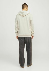 Sudadera beige claro con capucha ajustable, puños y dobladillo acanalados, combinada con pantalones de mezclilla oscuros y zapatos marrones. Textura de tela suave.