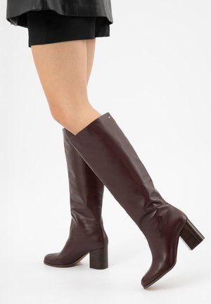 Beenzicht van een persoon die donkergroene, kniehoge leren laarzen met blokhakken en een zwarte rok draagt.