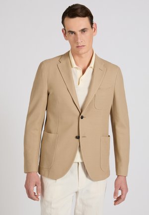 Uomo che indossa un blazer beige sopra una polo di colore chiaro e pantaloni bianchi, in piedi davanti a uno sfondo grigio chiaro e uniforme.