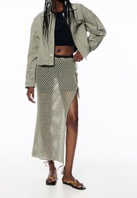 Kort spijkerjasje in olijfgroen over een zwart topje, gecombineerd met een lange, mesh-gepatteerde gebreide rok met een zijsplit en koordjes. Platte sandalen.