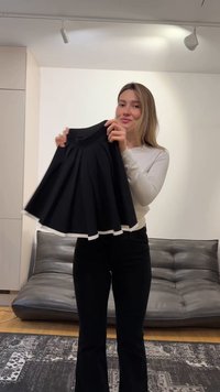 Giovane donna in piedi in casa che tiene una gonna nera con rifiniture bianche davanti a un divano grigio scuro e un tappeto decorato.