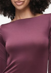 Femme aux cheveux bouclés portant un haut violet ajusté à manches longues et épaules dénudées, laissant voir son cou et une partie de son visage.