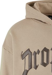 Beige Kapuzenpullover mit einem großen, dunkelbraunen grafischen Text auf der Vorderseite. Der Stoff wirkt weich, mit einem einfachen, lässigen Design und einer gemütlichen Kapuze.