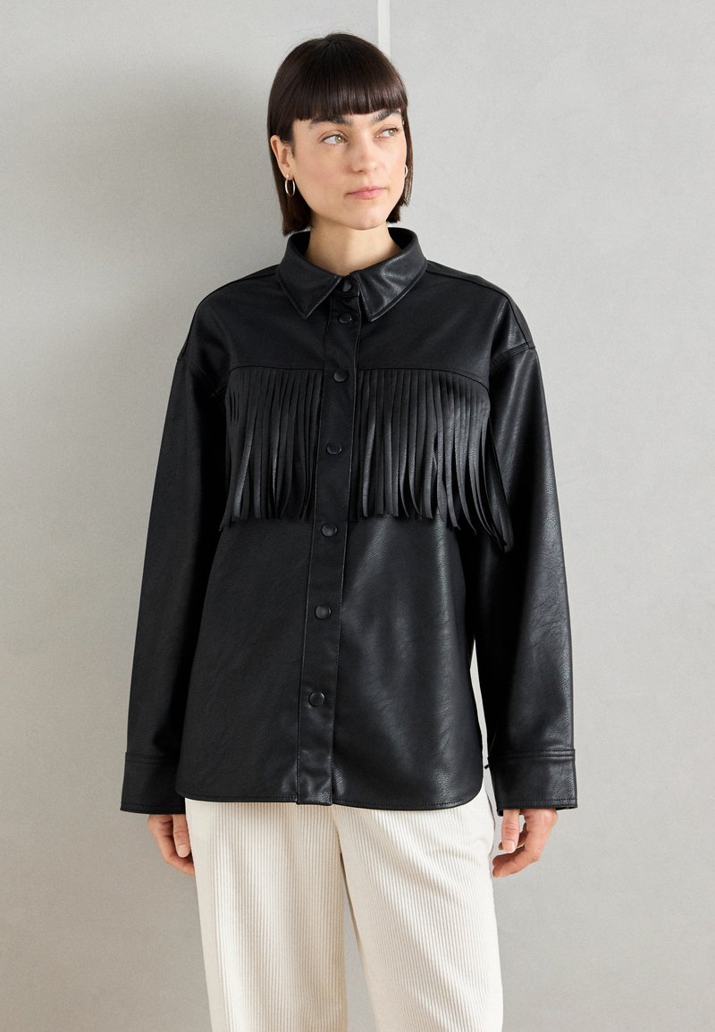 Chemisier en simili cuir noir avec des franges sur la poitrine, fermeture à boutons et manches longues. Texture lisse avec un col.