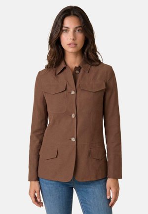 Donna con lunghi capelli scuri che indossa una giacca marrone con bottoni e quattro tasche con patta e jeans blu su uno sfondo semplice.