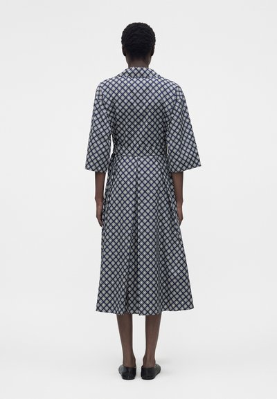 Άτομο που στέκεται με την πλάτη στην κάμερα, φοράει ένα μεσαίου μήκους μπλε φόρεμα με σχέδιο, φαρδιά μανίκια και μαύρα μπαλαρίνια σε λευκό φόντο.