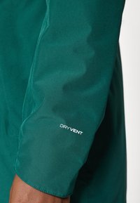 Grønt, vanntett stoff med en glatt tekstur. Har en synlig "DRYVENT"-logo på mansjetten. Rett skjær design på ermet.