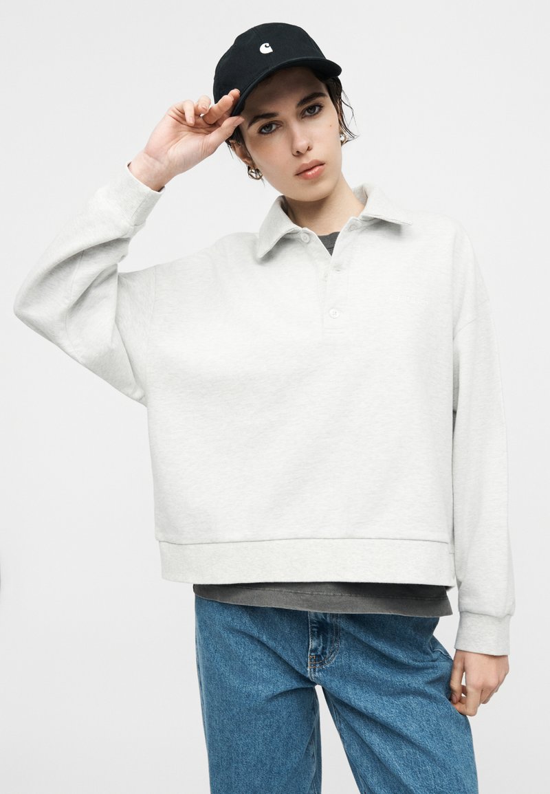 Joven con un suéter holgado de color gris claro, vaqueros azules y gorra negra, sujetando el borde de la gorra con la mano derecha contra un fondo liso.